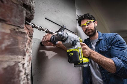 Man använder grön och svart borrmaskin för att borra genom betongvägg, med skyddsglasögon på.