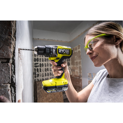 Kvinna med skyddsglasögon borrar i en vägg med en grön trådlös borrmaskin märkt "Ryobi".