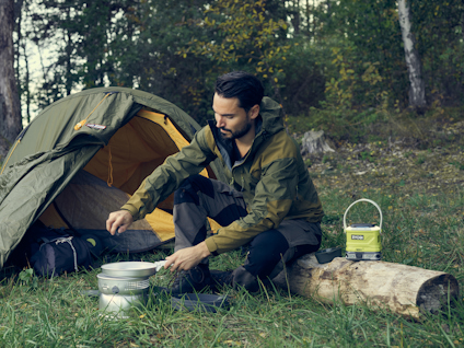 En person sitter utanför ett tält i skogen och lagar mat på en portabel spis.