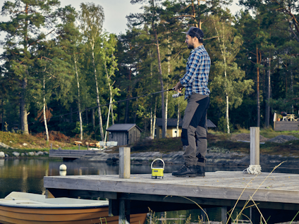 En person fiskar på en brygga vid en sjö, omgiven av träd och båtar.