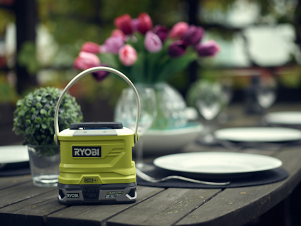 Grön bärbar Ryobi-lampa placerad på träbord med vita tallrikar och glasvas med rosa tulpaner i bakgrunden.