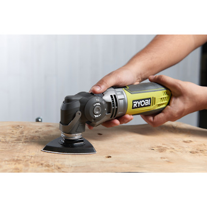 En elektrisk verktyg som hålls av två händer, används på en träyta, märkt "RYOBI".