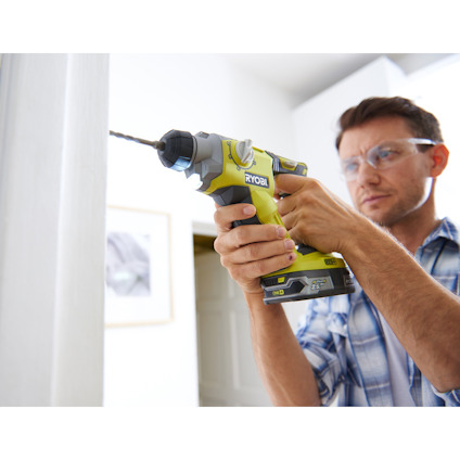 Person med skyddsglasögon använder en grön och svart Ryobi borrmaskin på en vit vägg.