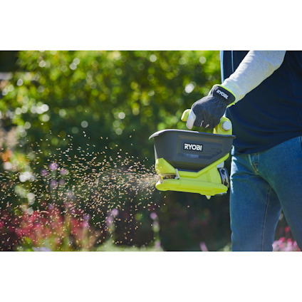 Person sprider frön i trädgården med en grön och svart Ryobi spridare.