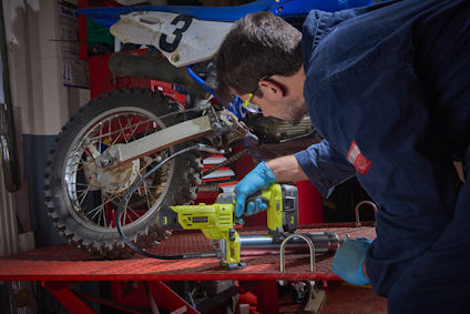 Person i blå overall använder en grön RYOBI smörjspruta nära bakhjulet på en motorcykel.