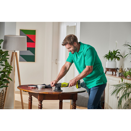 En person använder en grön borrmaskin vid ett runt bord i ett vardagsrum med växter och konst på väggen.