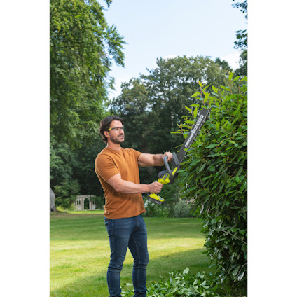 Person klipper en högre buske med en batteridriven häcksax på en gräsmatta.