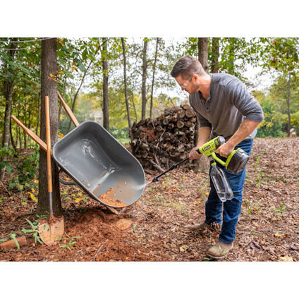 Man with a drill emptying soil from a wheelbarrow supported by trees in a wooded area.