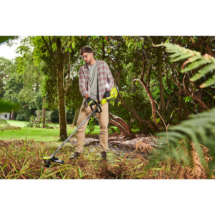 Man använder en grön och svart grästrimmer i en trädgård med gröna växter och träd.