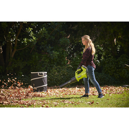 Person använder en lövblåsare utomhus, omgiven av löv och ett trädgårdsskräppåse.