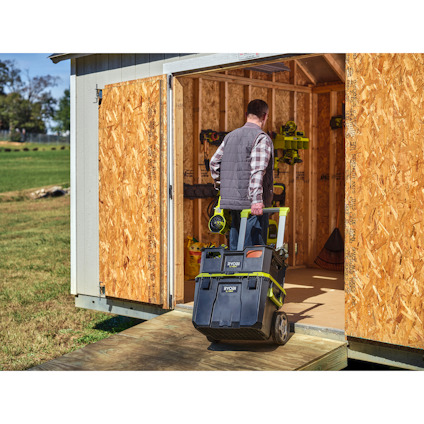 Man pushing a stack of toolboxes on wheels into a wooden shed with open doors.