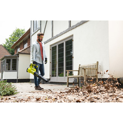 Person använder en lövblåsare för att blåsa bort löv från en uteplats utanför ett hus.