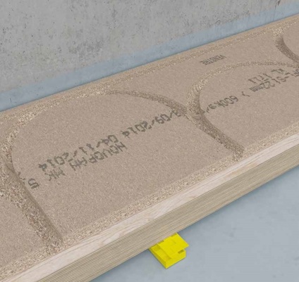 Träfärgad spånskiva med halvcirkulära förfrästa spår och text, placerad mot en betongvägg ovanpå en gul stödstruktur.