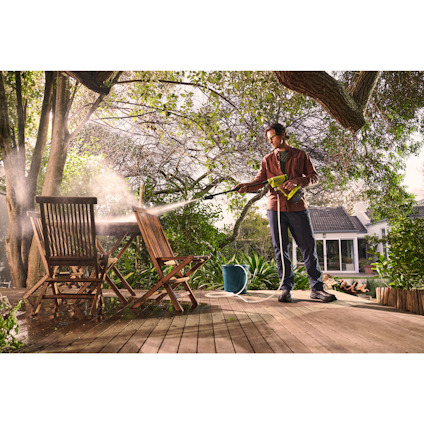 Person spolar av två trästolar med en mellantryckstvätt av märket RYOBI på en träveranda omgiven av träd och en trädgård.