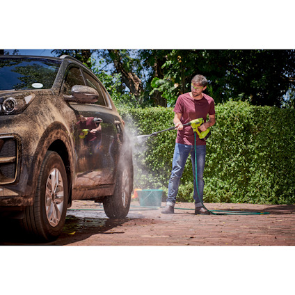 Man i röd tröja och jeans tvättar en lerig bil med en mellantryckstvätt av märket Ryobi på en stengång omgiven av grönska.