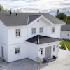 Tvåvånings hus med vita träfasader, svarta takpannor och en stensatt uppfart. Omgivande grönska och molnig himmel.