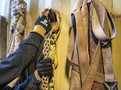 Hand med handske greppar rostig kedja bredvid upphängda bälten och rep mot beige vägg.