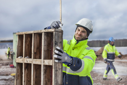 Person i hjälm och reflekterande jacka använder en måttstock för att mäta träform medan andra arbetar i bakgrunden.