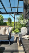 Inglasad uteplats med grå soffa, kuddar, bord, krukväxter och utsikt mot grönska och blå himmel.