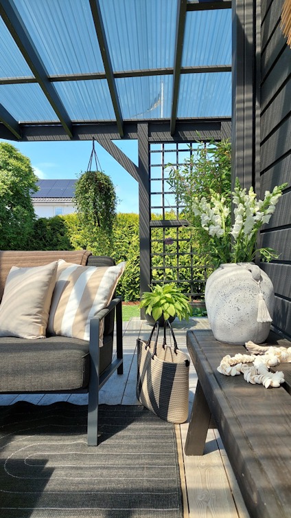 Inglasad uteplats med grå soffa, kuddar, bord, krukväxter och utsikt mot grönska och blå himmel.