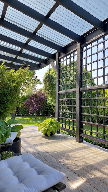 Pergola med träribbor och tak av glas, omgiven av grönska och blommor i trädgårdsmiljö.