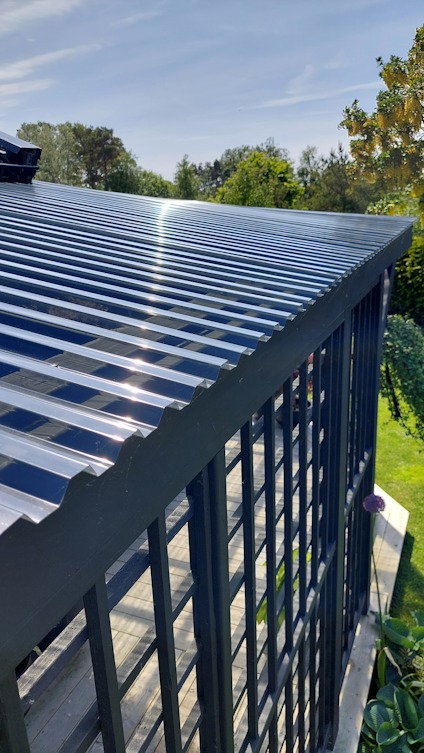 Svart pergola med genomskinligt tak av korrugerad plast, omgiven av grönska och blå himmel.