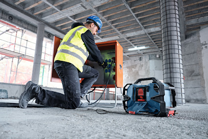 Person i byggarbetskläder arbetar med en elektrisk utrustning framför en orange skåp i en ofärdig byggnad.